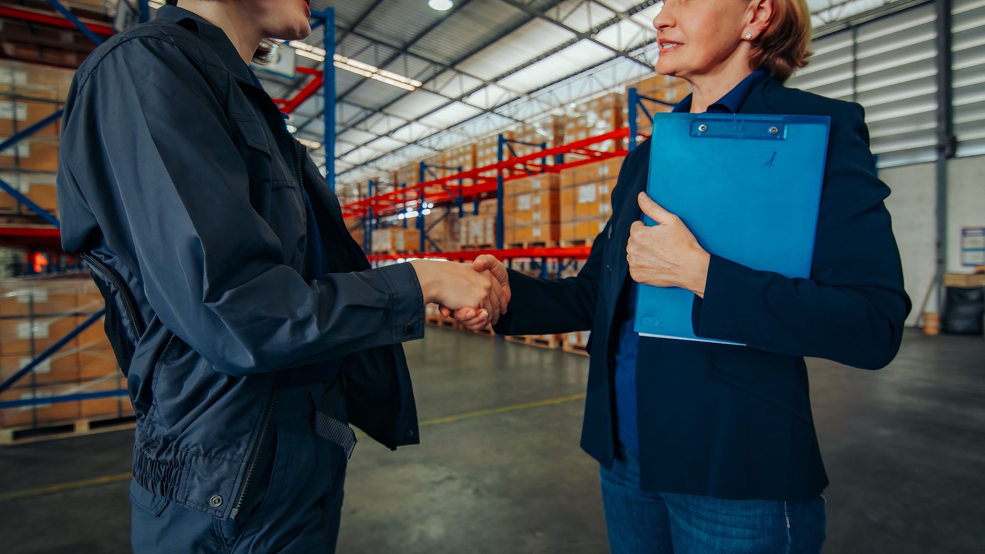 Two logistics experts shake hands in agreement inside a warehouse, signifying a successful negotiation or partnership.